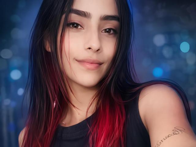 Tatis69 ha lunghi capelli neri con punte rosse, occhi marroni e un tatuaggio sul braccio destro. Indossa una canotta nera e sorride dolcemente..
