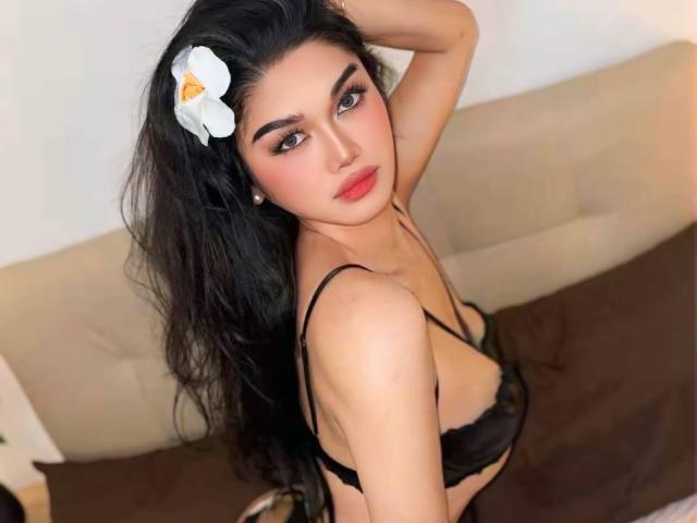 MiacoCoa with long black hair, a white flower in her hair, wearing a black bra, looking up with a sexy, confident expression, showing her tits.