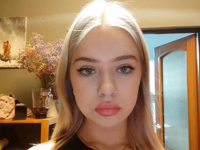 BlossomRoeder has blonde hair, green eyes, and pink lips. Her makeup includes black eyeliner. She's wearing a black top, with a wooden door and floral vase in the background.