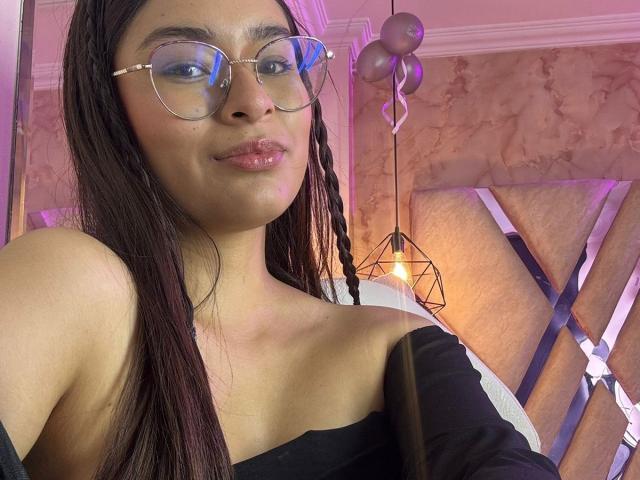 IsaDoux with shiny glasses, dark braids, glossy pink lips, and a black off-shoulder top, showing her bare shoulders and collarbone, smiling subtly.
