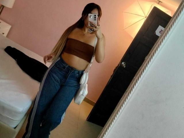 ZoeyClark with long blonde and brown hair, wearing a brown tube top and high-waisted blue track pants, holding a phone, mirror selfie, showing her ass and a white bag.