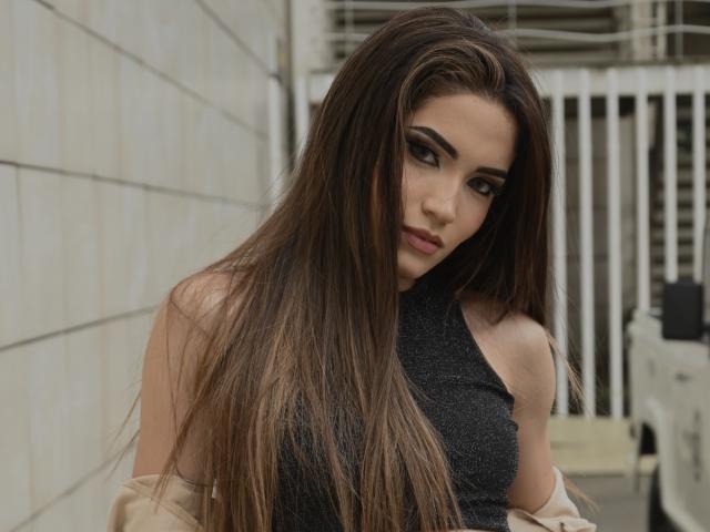 TaylHell has long brown hair, dark eye makeup, and a serious expression. She's wearing a sleeveless black top with her tan jacket off. Boobs partially visible.