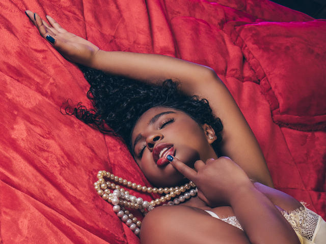 LisaMendez with dark curly hair, red lips, and blue nails, lying on red fabric, touching her tongue to a pearl necklace, wearing a lace top, arms raised.