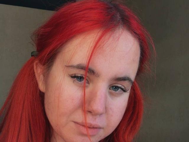 Close-up of CaseyChase with bright red hair in pigtails. She has fair skin with a few small blemishes and dark, slightly arched eyebrows. Her blue eyes are slightly downcast, and she has long, dark eyelashes. Her expression is neutral, with a slight, closed-lip smile. The texture of her hair is slightly messy, with strands falling over her face. The background is out of focus, emphasizing her facial features. The image is sharp, with natural lighting highlighting her skin and hair.