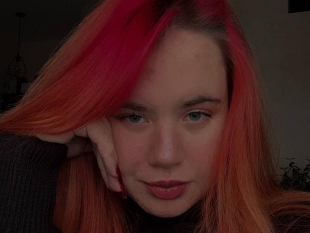 Close-up of CaseyChase with a serious expression. CaseyChase has long, straight, vibrant red-to-orange gradient hair, with the red on the left side and orange on the right. She's resting her head on her right hand, which has red-painted nails. Her blue-green eyes are slightly downcast, and she has pinkish freckles on her cheeks. She's wearing a dark, ribbed sweater. The lighting is soft, creating subtle shadows on her face. The background is blurred and dark, with a small, indistinct plant visible on the right. Her lips are slightly parted, showing a subtle smile.