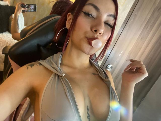 ValeryHarllow with red hair, hoop earrings, and a grey zip-up top, blowing a bubble, showing her tits, tattoos on her shoulder, taking a selfie in a chair.