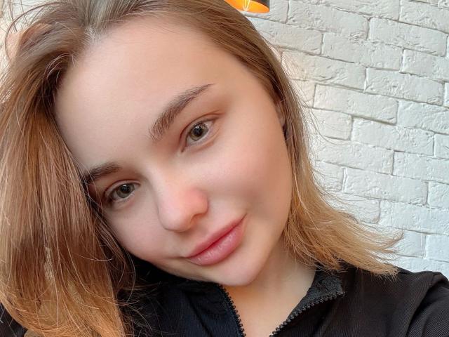 IvyEvans-hot has light brown hair, green eyes, and a slight smile. She's wearing a black zip-up and her nose is small. Her skin is fair and she looks cute.