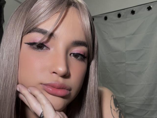 KateGoddes with pink eyeshadow, black winged eyeliner, and pink lipstick, has her chin on her hand, showing a tattoo on her arm, with long, straight, blonde hair.