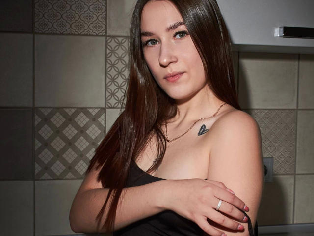 SweetCookies-hot with long brown hair, crossed arms, black strapless top, small heart tattoo on chest, subtle smile, and red nail polish, wearing a thin gold necklace.