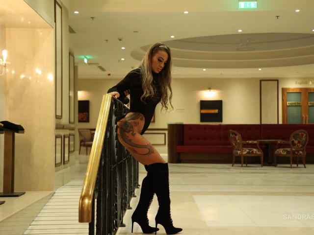 SandraSayd with long blonde hair, black top, black knee-high boots, and tattoos on her thigh, leaning on a gold railing, showing her curvy ass.