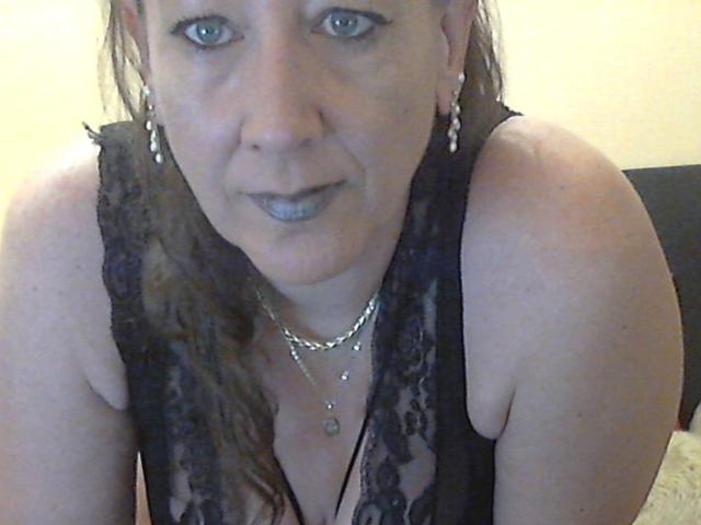 XMyriamX with gray lipstick, wearing black lace top, showing her boobs, earrings, and necklace, looking into the camera with a slight smile. Her hair is wet and brown.