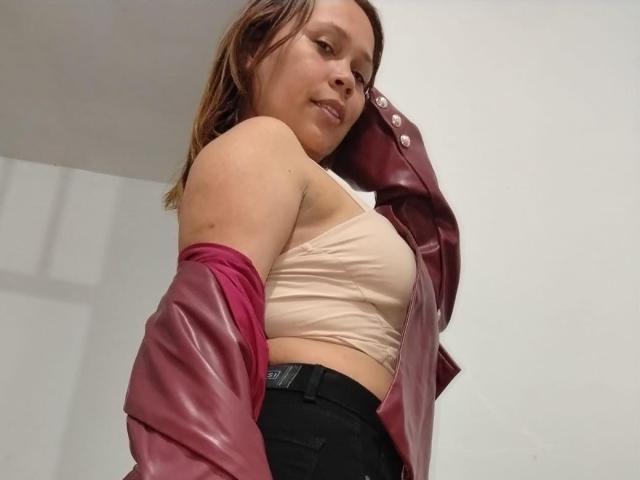 SophiaQueens with pink leather jacket draped over her shoulders, beige bra, and black pants, smiling down at the camera, showing her tits. Her brown hair is loose.