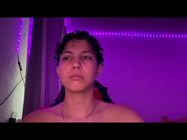 Shirtless AlinaRicker with curly dark hair and a slight scab on the forehead, wearing a silver necklace, under purple LED lights. Face looks serious, slightly upturned nose.