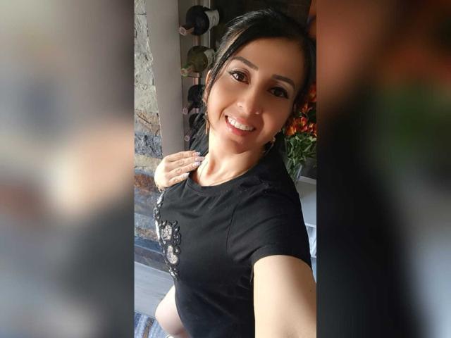 Briananice with dark hair, smiling, wearing a black shirt, holding phone for selfie, visible boobs, hand on chest, blurred foreground, indoor setting, casual, cute, attractive, brown eyes.
