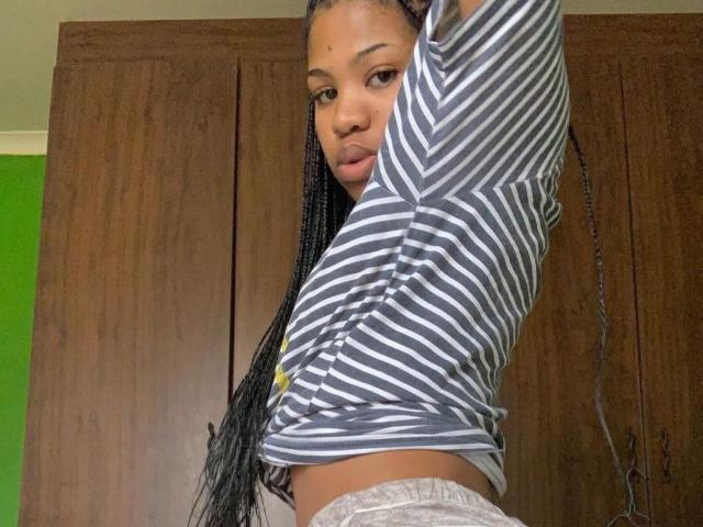 Mbebo with long braids, striped shirt, and gray pants, lifting shirt to show a sliver of her tan, flat ass. Brown wooden doors in the background.