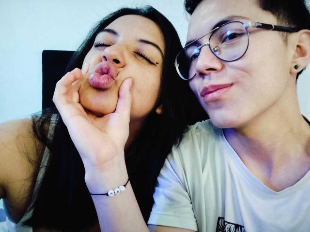 TinnaNCharlie with dark hair, pouty lips, and a nose piercing, gets her face pinched by TinnaNCharlie with glasses and short hair, wearing a white tee.