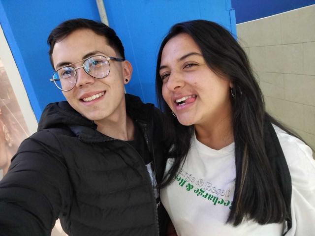 TinnaNCharlie with short black hair and glasses, wearing a black puffy vest, stands next to TinnaNCharlie with long black hair, wearing a white shirt, both smiling.
