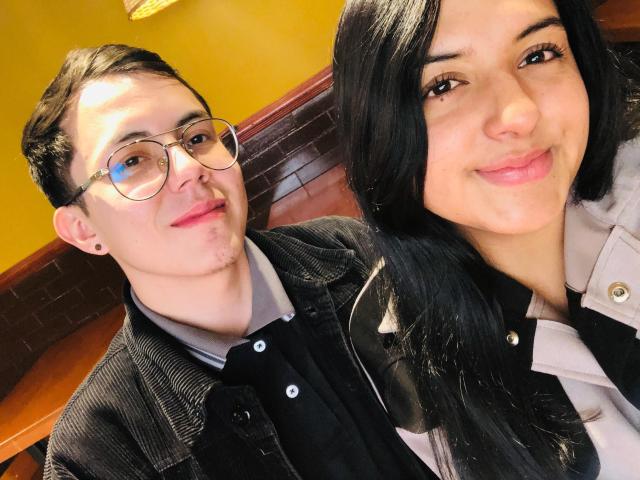 TinnaNCharlie with glasses, short black hair, and a slight mustache, wearing a black shirt and pinstripe jacket, next to TinnaNCharlie with long black hair, pink lips, and a smile.