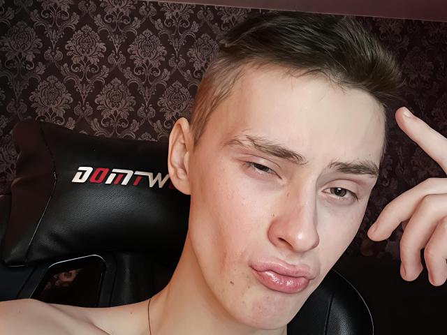 HomerBart with short, brown hair, pouty lips, and a slight smirk, making a "shh" gesture with their finger. Black leather chair with "AWW" logo in background.