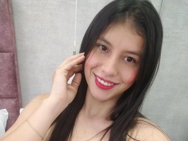 MadissonFontaine has long black hair, pink blush, and bright pink lipstick. She's smiling with red lips, touching her ear, and wearing a gold bracelet. She's a cute bitch.