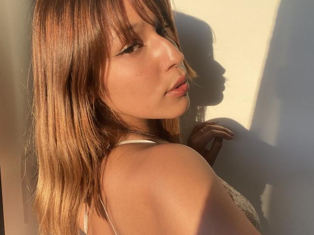 KiaraHernandezX with medium brown hair, looking over her shoulder, sunlight on her face, slight smile, wearing a white lace top, hand near her neck, shadow on wall.