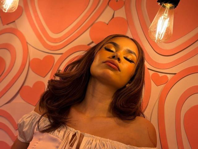 MargariitaZa with closed eyes, wavy brown hair, and a relaxed expression, wearing a white off-shoulder top, under a hanging light bulb, showing a calm, sexy vibe.