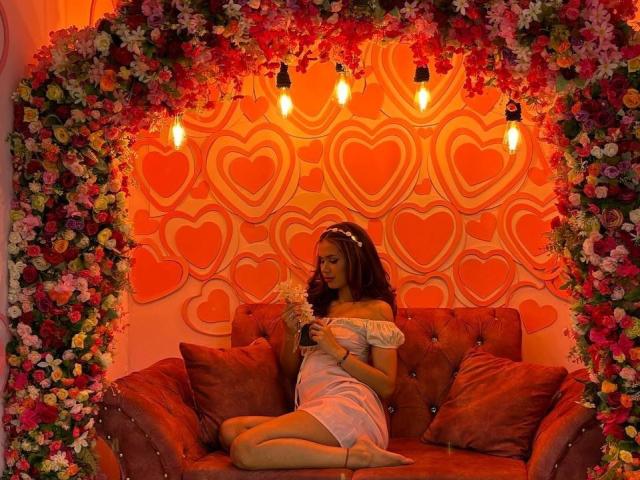 MargariitaZa with wavy brown hair, off-shoulder white dress, and flower crown, sitting on red velvet couch, holding flowers, surrounded by floral arch, heart-patterned wall, and exposed bulbs.