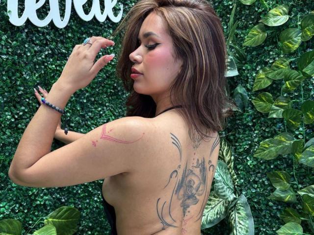 MargariitaZa with brown hair, pink blush, and tattoos on her back and arm, showing her tattoos and a ring, topless, wearing a black strap, looking to the side.