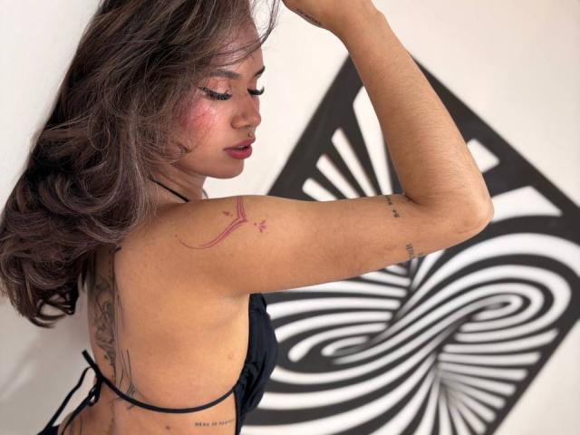 MargariitaZa with brown wavy hair, black bikini, red lipstick, tattoos on arm and back, raised arm, white wall with black abstract pattern, slightly turned side profile.