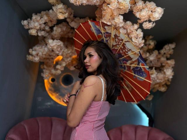 MargariitaZa with dark wavy hair, pink checkered dress, holding red and gold umbrella, showing tattoo on left shoulder, standing in front of pink tufted sofa.