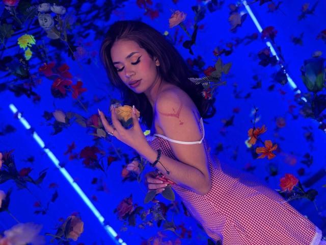 MargariitaZa with long brown hair, wearing a red-and-white checkered dress, holding a yellow rose, has a red scar on her right shoulder, against a blue, flower-filled background.