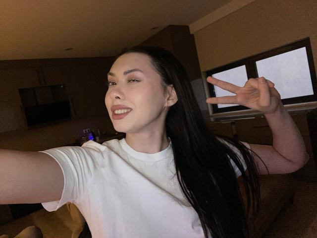 GwenOnline with long black hair, white t-shirt, fair skin, smiling, making a peace sign, in a dimly lit room, showing off her cute face and slightly parted pink lips.