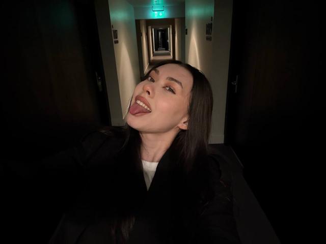 GwenOnline with long brown hair, tongue out, sticking it to her upper lip, wearing a black jacket, in a dimly lit hallway, looking directly at the camera.
