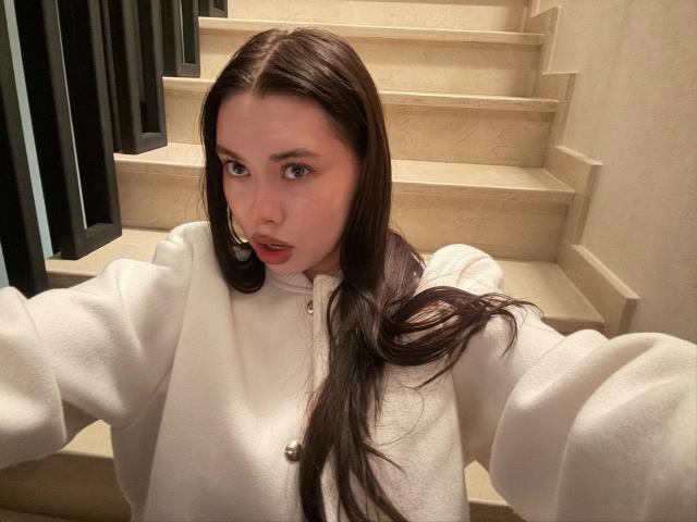 GwenOnline with long brown hair, wearing a white fluffy sweater, taking a selfie on a staircase. She has a small nose ring and a surprised expression on her face.