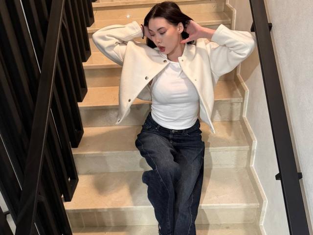 GwenOnline sitting on stairs, arms behind head, wearing a white jacket, white shirt, and dark blue jeans, with her mouth slightly open, looking relaxed.