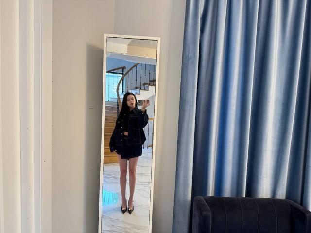 A full-length mirror reflects GwenOnline standing in a room. She has long dark hair, wears a black oversized jacket and short black skirt, and black high heels. Her right hand is raised, and she looks at the mirror. The background shows a staircase with wooden steps and a glass railing. Blue curtains are on the right, and a dark gray armchair is partially visible. The floor is white marble. The mirror has a white frame. The image is clear and well-lit.