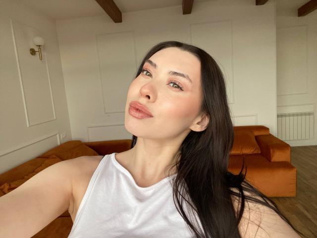 GwenOnline has pale skin, long black hair, and is wearing a white tank top. She's pouting with pink lips, and has small tits visible. She's taking a selfie.