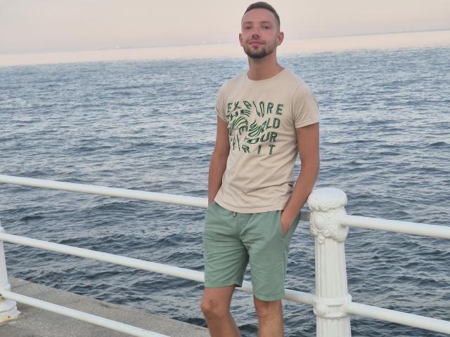Cocco69 stands with hands in green shorts, beige shirt, and short beard, looking at camera. Ocean and white railing in background. Hands in pockets, casual, relaxed.