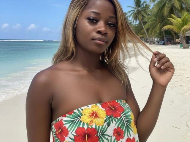 EbonyAngel-hot with dark skin and blonde hair, wearing a strapless top with red and yellow hibiscus flowers, standing on a beach, holding her hair with a slight smile.