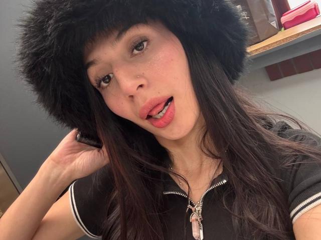 SophieBeaumont with black fur hat, long dark hair, slightly open mouth, nose piercing, black zip-up shirt, right hand in hat, pink lips, light skin, brown eyes.