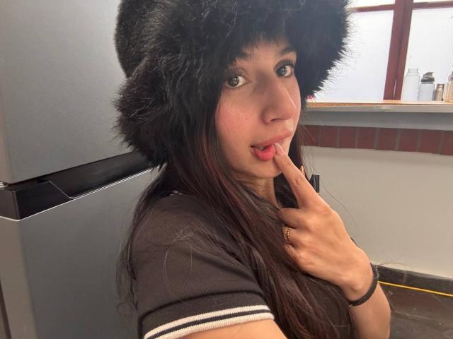 SophieBeaumont with long dark hair, wearing a black furry hat, touching her pink-tipped tongue with her finger, wearing a brown shirt, in a kitchen-like setting.