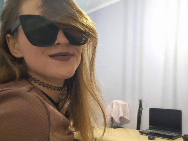 Millfka with long blonde hair, black sunglasses, dark lipstick, choker, brown ribbed shirt, smiling, laptop on desk, guitar, and pink towel in the background.