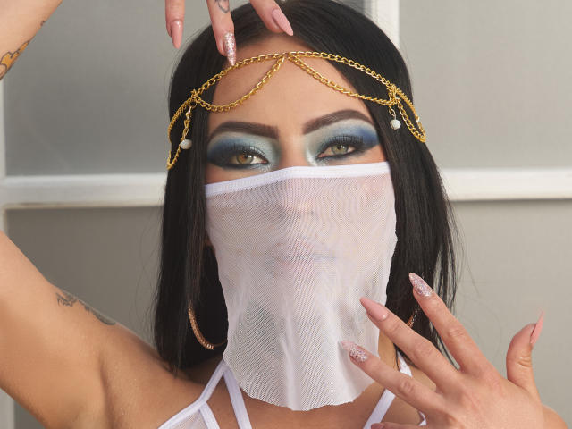 LunaBaster with black hair, blue-green eye makeup, gold chain headpiece, white mask covering nose and mouth, long pink nails, visible tattoos, white halter top, indoors.