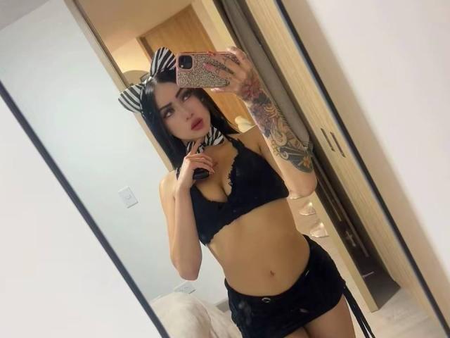LunaBaster with black bra, black shorts, zebra bow, and tattooed arm, taking a mirror selfie, showing her tits and ass, holding a glittery phone.