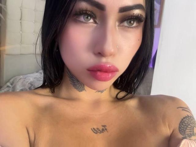 LunaBaster with black hair, pink lips, tattoos on neck and chest, and visible collarbone, has a nose ring and is topless, showing her tits and small tattoos.