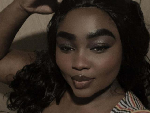 ReinaEbanox with dark skin, thick eyebrows, purple eyeshadow, long curly black hair, and full lips, lying down with a hand raised, wearing a striped top.
