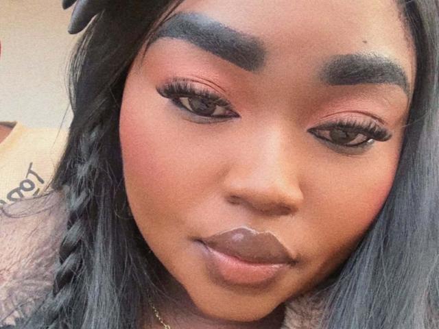 ReinaEbanox's face with dark brown skin, thick black eyebrows, pink eyeshadow, long eyelashes, and brown lipstick. She has black hair in a braid and a gold necklace.