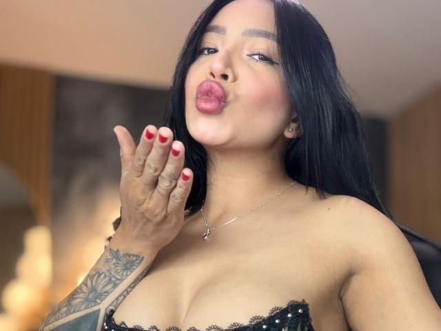 Abie69 with black hair, pouty pink lips, red nail polish, tattooed arm, and large boobs, kissing air, wearing black lace strap, silver necklace, in a slightly blurred photo.