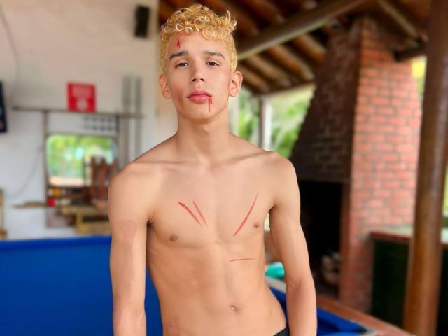 Shirtless ArdienteCouple with curly blonde hair, light skin, and red scratches on chest and arms, standing in blue-walled room. ArdienteCouple has a slight, cute, and sexy look.