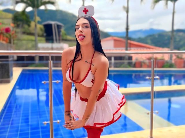 ArdienteCouple with long black hair, tongue out, wearing a red and white nurse outfit, showing big boobs, tattoos, and a hat, bending over by a blue-tiled pool.